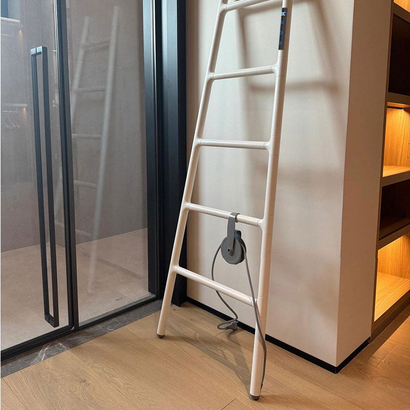 A real-life interior shot of a cream white ladder-style electric heated towel rack leaning against a neutral wall between a sleek black-framed glass sliding door and a wooden walk-in closet with warm built-in lighting.