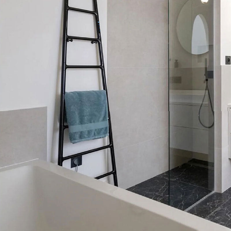 A realistic modern bathroom installation showing a matte black ladder-style electric heated towel rack leaning against a white wall with a teal towel, next to a freestanding bathtub and a glass shower enclosure with black marble flooring
