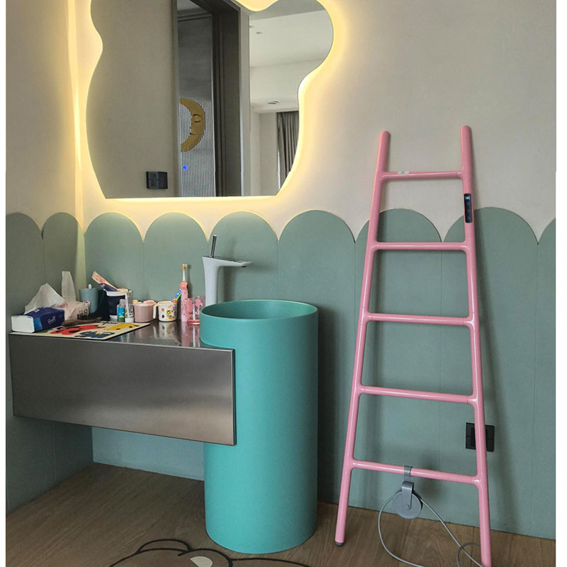 A lively real-life kids' bathroom installation featuring a pastel pink ladder-style electric heated towel rack leaning against a sage green scalloped panel wall next to a mint green pedestal sink and a backlit wavy mirror.