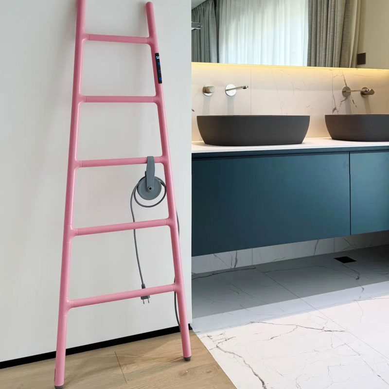 A real-life bathroom scene featuring a soft pink ladder-style electric heated towel rack leaning against a white wall next to a luxury modern vanity with double dark stone basins and a navy blue floating cabinet