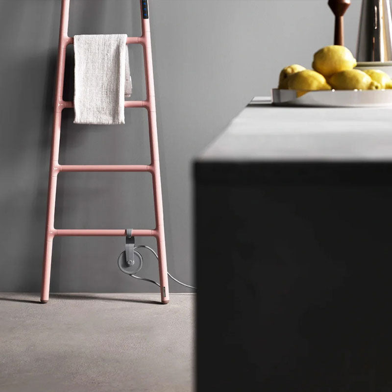Elegant pastel pink ladder-style electric heated towel warmer with a smart touch display and grey cord storage wheel, leaning against a grey wall in a modern kitchen or living area next to a bowl of fresh lemons.