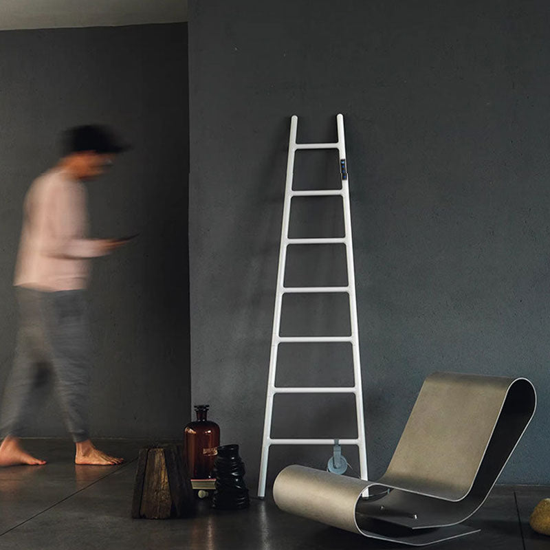 Editorial lifestyle shot of a white ladder-style electric towel rack leaning against a dark textured concrete wall in an artistic, industrial interior featuring a modern designer lounge chair.